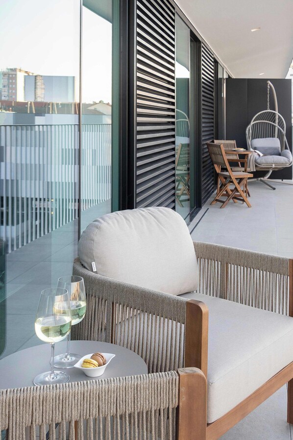 A balcony with wicker furniture, a glass table with a wine glass and snacks, and a view of the cityscape.