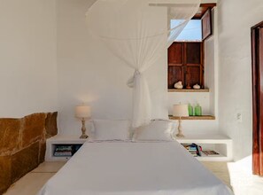 Un dormitorio con cama, mesitas de noche, una ventana con contraventanas de madera y un estante con artículos de aseo.
