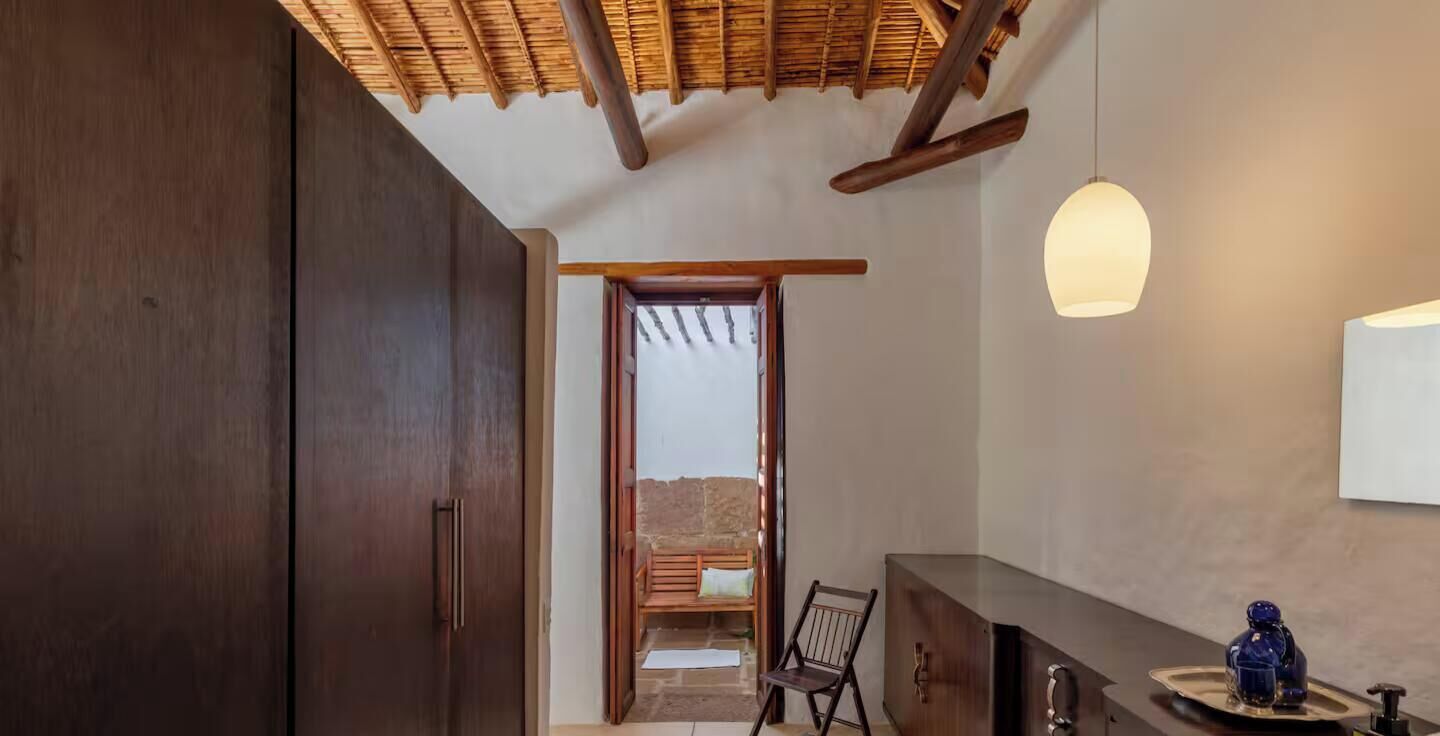 Una habitación con techo de madera, un banco, una silla, un armario y una lámpara colgante.