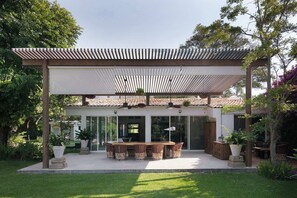 Eine überdachte Terrasse mit Esstisch und Stühlen, umgeben von Pflanzen und einem Gebäude mit großen Fenstern.