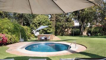 Une piscine extérieure avec un espace patio, entourée de pelouse et d’arbres.
