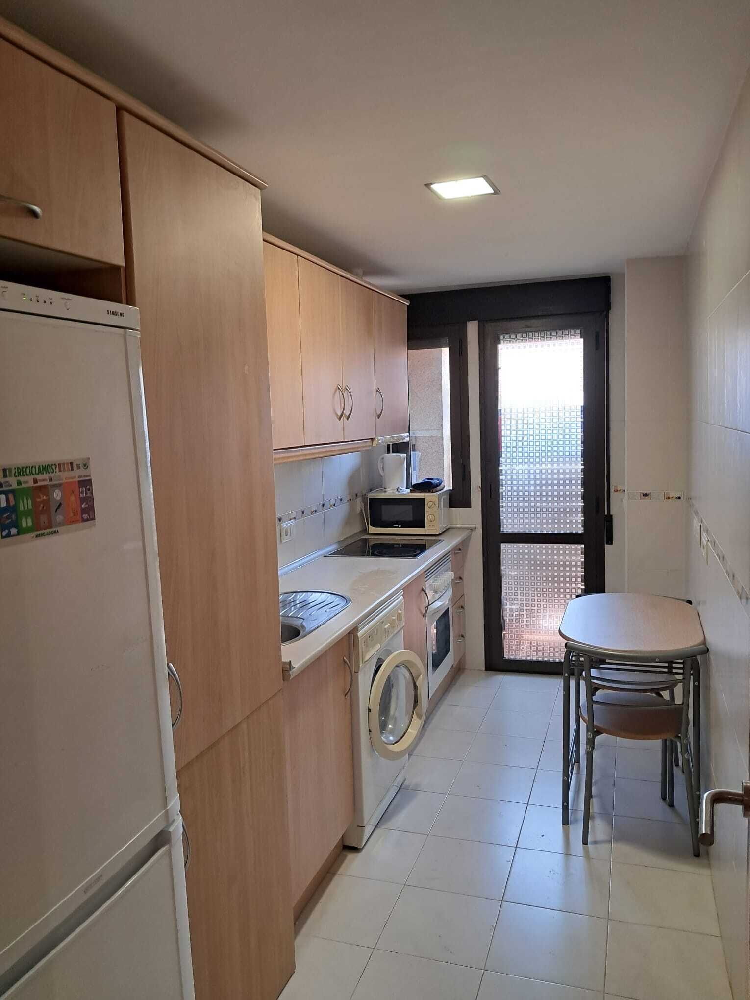 Cocina compacta con armarios de madera, refrigerador, microondas, lavadora y una pequeña mesa de comedor con sillas.