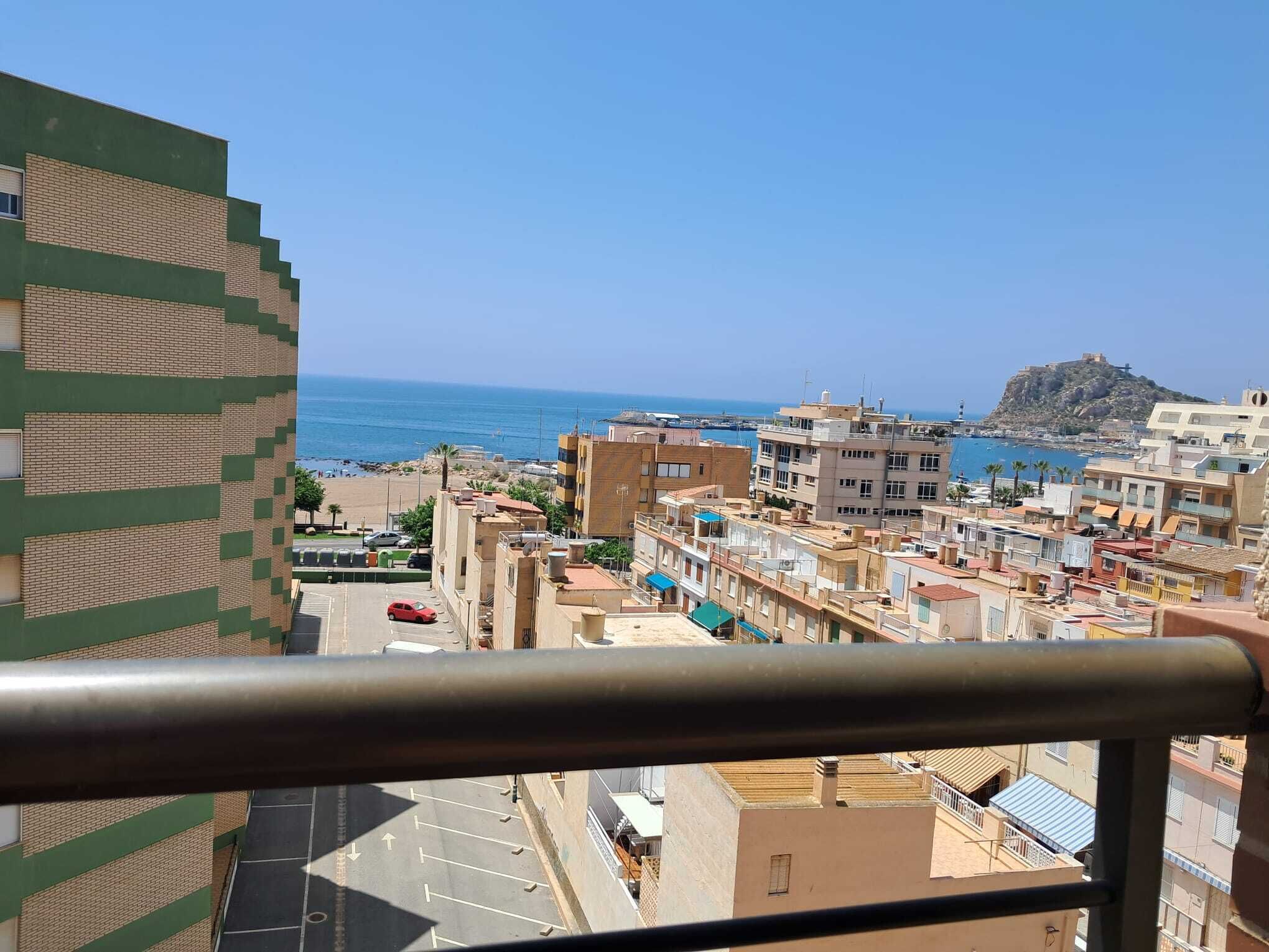 Un paisaje urbano costero con edificios, una playa y una colina con una fortaleza.