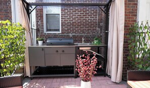 An outdoor kitchen area with a grill, sink, and storage cabinets, surrounded by potted plants and a brick wall.