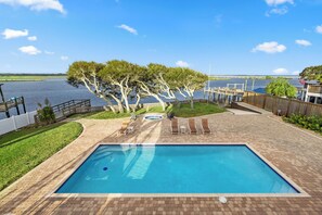 A rectangular swimming pool with a tiled edge, surrounded by a brick deck, and overlooking a body of water with a dock and trees. - Luxury Waterfront Retreat - Pool, Hot Tub & Breathtaking Sunsets - Sleeps 12 (St. Augustine)