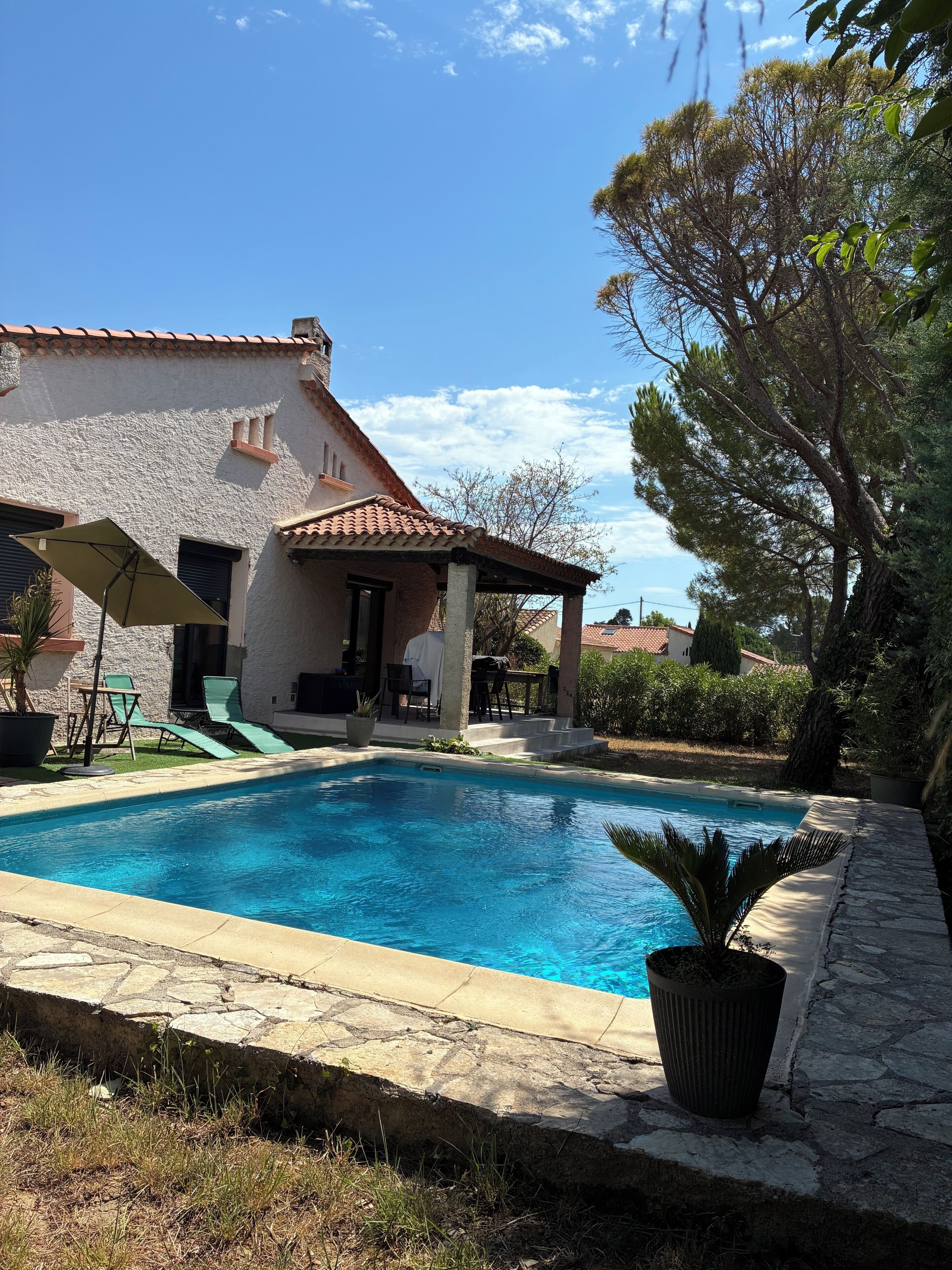 A swimming pool with a stone border, a covered patio with outdoor furniture, and a building with a tiled roof.