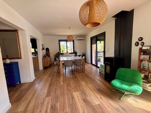 Uma sala ampla com uma mesa de jantar, cadeiras, uma poltrona verde, lareira e uma grande janela.