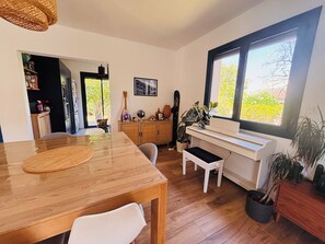 Uma área de jantar com mesa e cadeiras de madeira, um piano e uma janela com vista para o verde.
