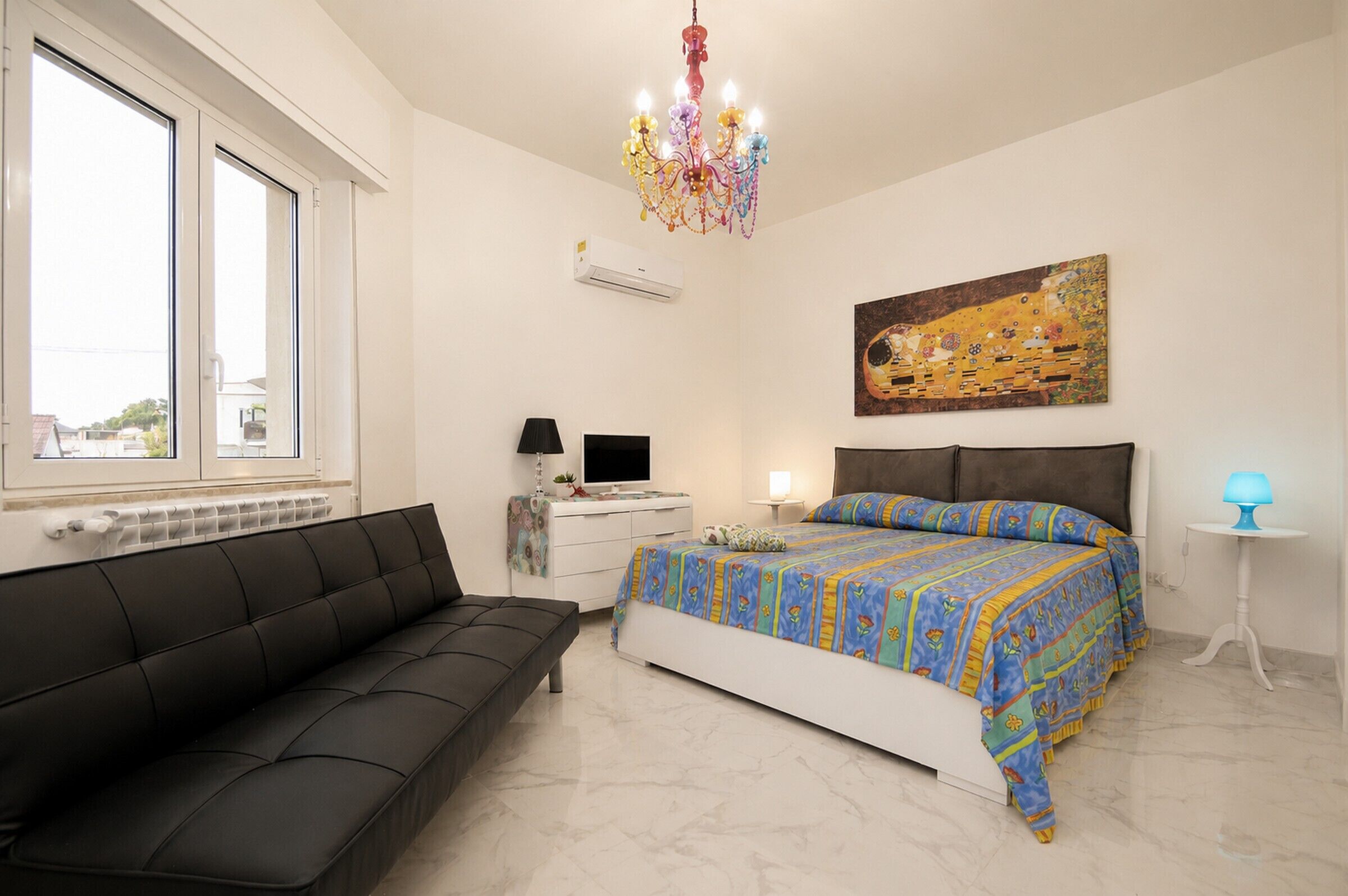 Un dormitorio con cama, sofá negro, escritorio con computadora, un cuadro en la pared y una araña.