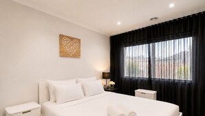 Habitación de hotel con una cama grande, una mesita de noche con lámpara, un pequeño cubo de basura y una ventana con cortinas negras.