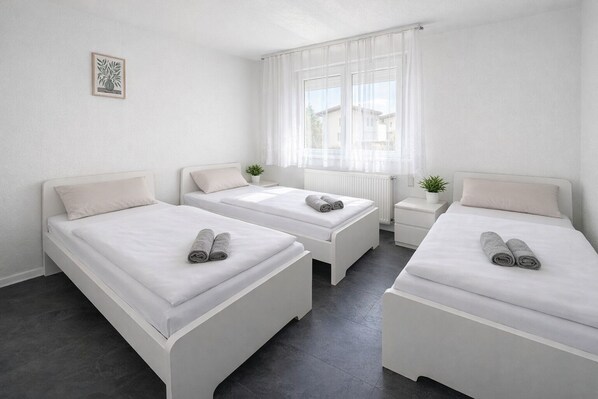 A room with three beds, a small bedside table with a plant, and a window with sheer curtains.