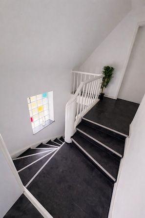 Una escalera con peldaños oscuros y barandillas blancas, un ventanal con diseño de cuadrícula y una planta en el rellano.