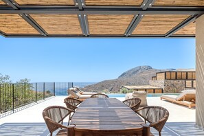 A covered outdoor seating area with a wooden table and wicker chairs, overlooking a mountainous landscape and a body of water. - Metatulia Villa 6 – Private Pool, Jacuzzi & Views (Plakias)