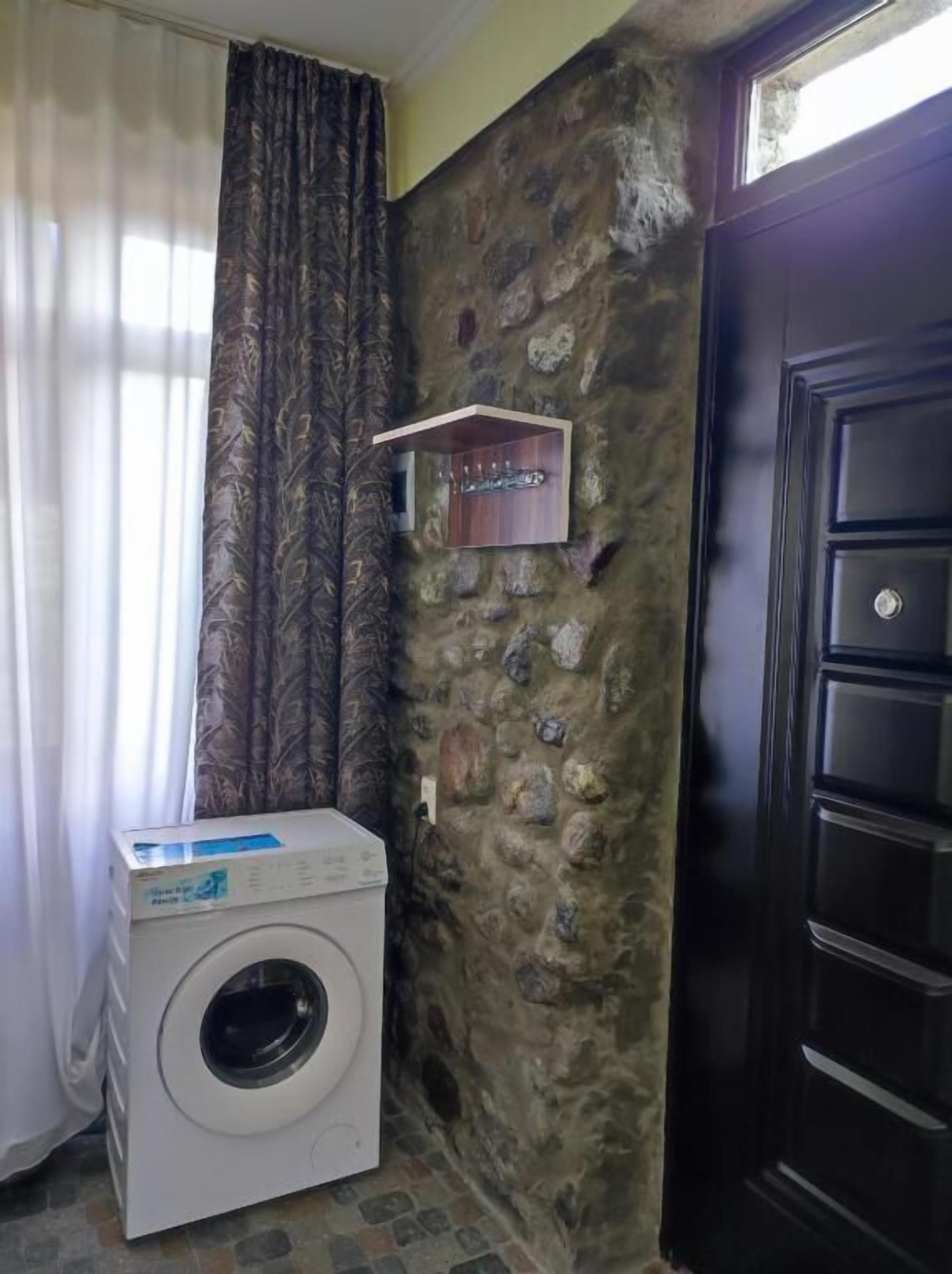 A laundry area with a washing machine, a stone wall, a door, and a window with curtains.