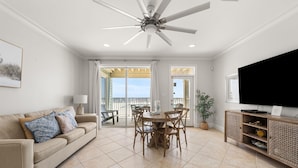 A spacious living room with a ceiling fan, a large flat-screen TV, a dining area with wooden chairs, and a balcony with a view. - The Hammock Dunes Collection (Orange Beach)