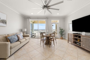 A spacious living room with a ceiling fan, a large flat-screen TV, a dining area with wooden chairs, and a balcony with a view. - The Hammock Dunes Collection (Orange Beach)