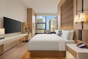 A modern hotel room with a large bed, a flat-screen TV, a desk, and a view of high-rise buildings.