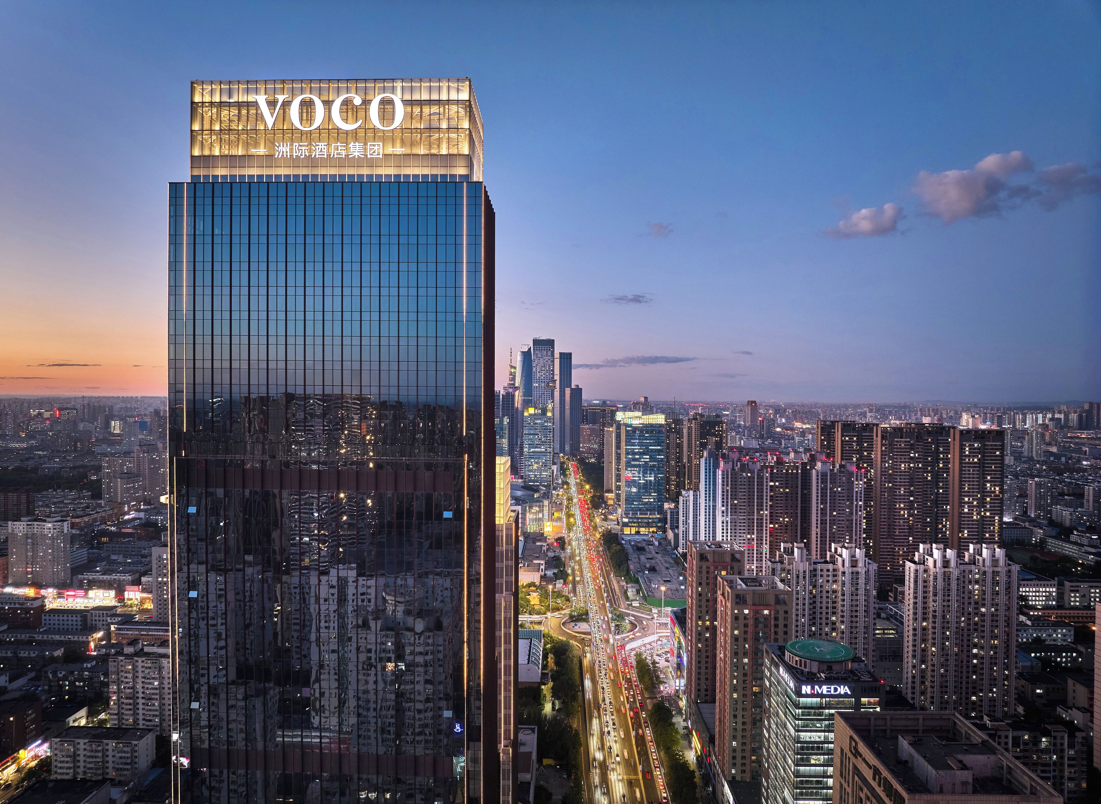 A high-rise building with a prominent 'Voco' sign, surrounded by a dense urban skyline with numerous other high-rise buildings.