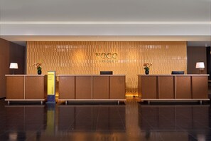 Un espace d’accueil avec un mur revêtu de panneaux en bois sur lequel figure le nom « VOCO », ainsi qu’un comptoir d’accueil équipé d’un ordinateur et agrémenté d’un vase de fleurs.