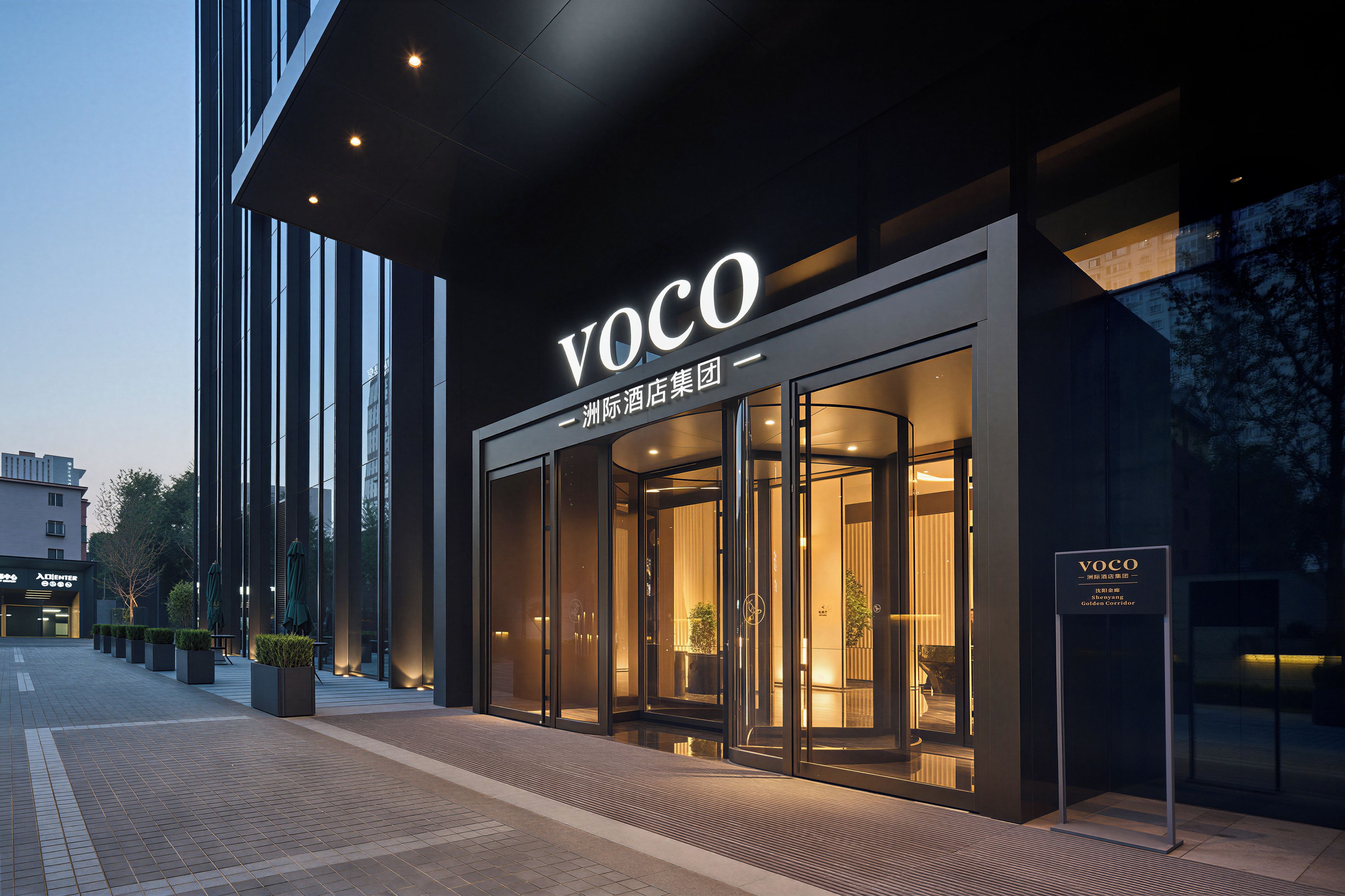 A modern hotel entrance with a glass facade, a prominent 'VOCO' sign, and a well-lit interior visible through the doors.