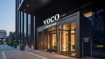 A modern hotel entrance with a glass facade, a prominent 'VOCO' sign, and a well-lit interior visible through the doors.