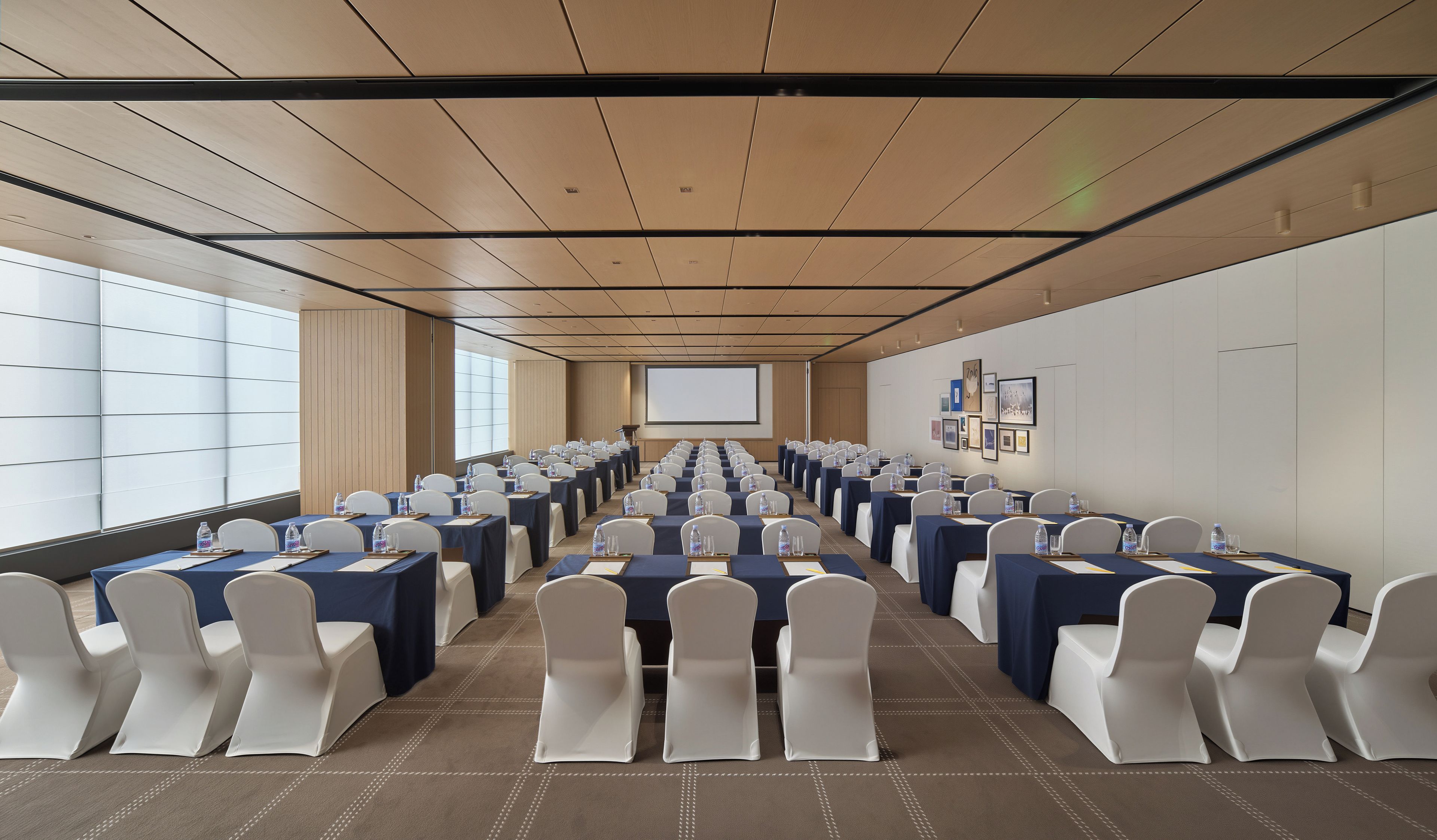 Ein Konferenzraum mit Tischen und Stühlen, einer Projektionsleinwand und gerahmten Bildern an der Wand.