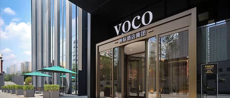 A modern building facade with a prominent 'VOCO' sign, large windows, and outdoor seating area with umbrellas.
