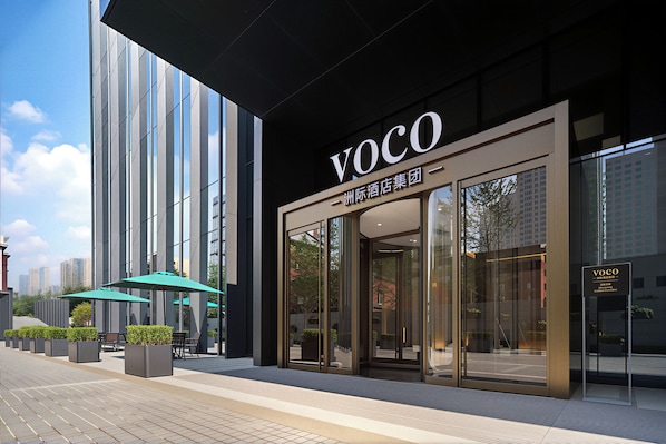 A modern building facade with a prominent 'VOCO' sign, large windows, and outdoor seating area with umbrellas.