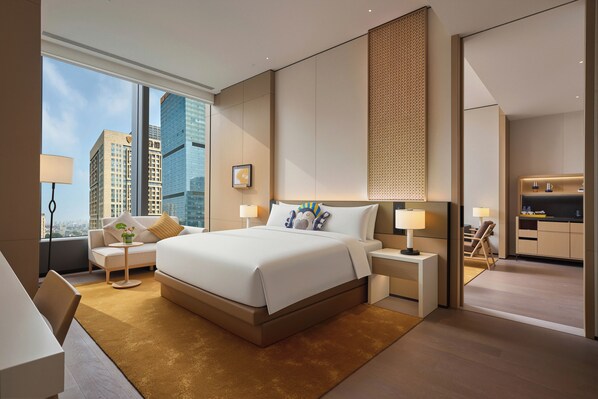 Une chambre d’hôtel moderne dotée d’un grand lit, d’un coin salon avec un canapé et un fauteuil, d’une petite table avec une lampe, et offrant une vue sur les toits de la ville grâce à de grandes fenêtres.