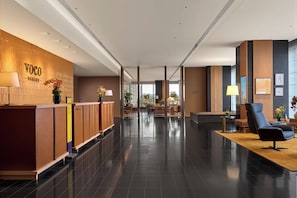 A modern hotel lobby with a reception desk, seating area, and a view of the cityscape through large windows.