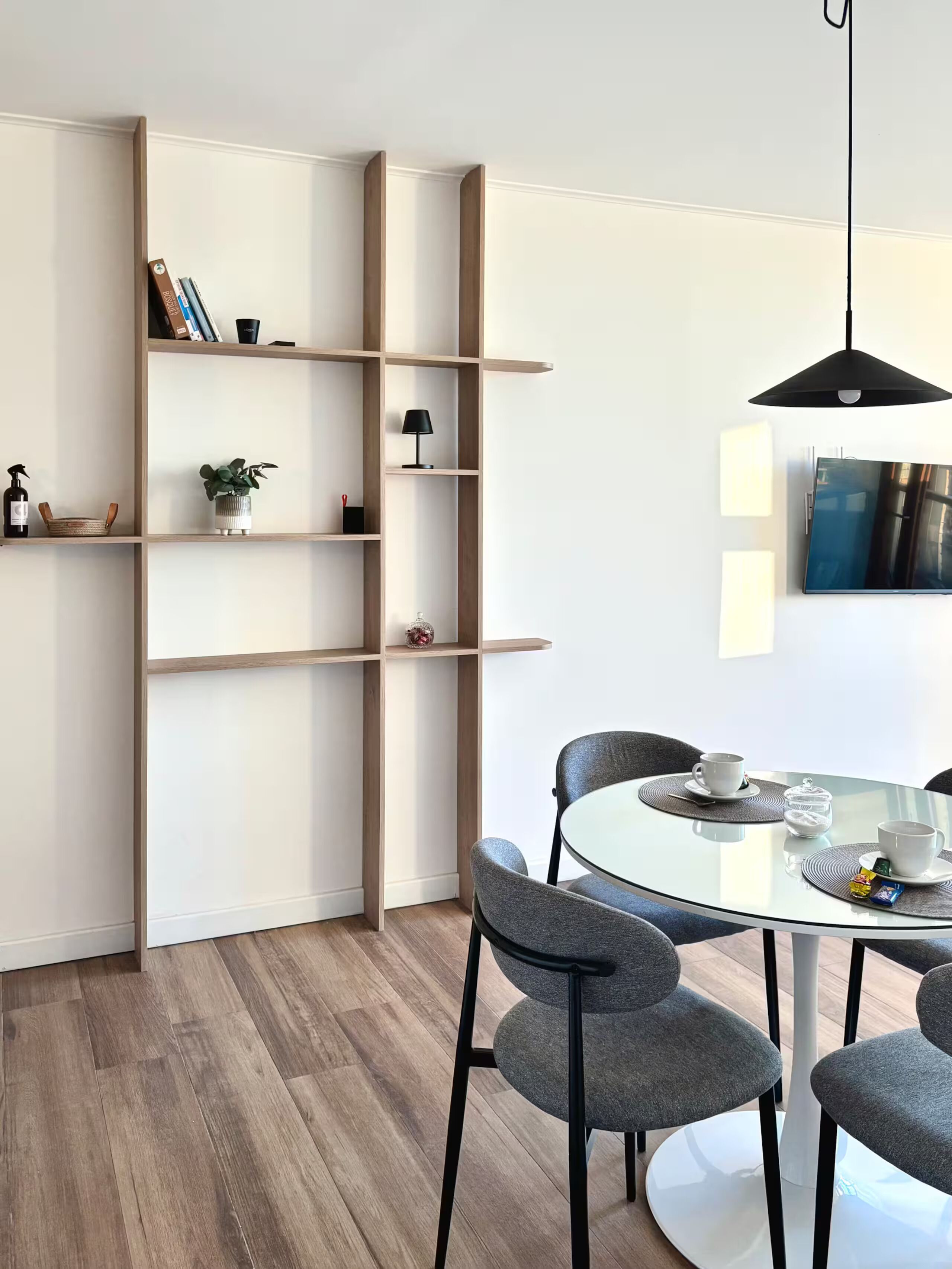 A modern dining area with a round glass table, grey upholstered chairs, a wooden bookshelf, and a black pendant light.