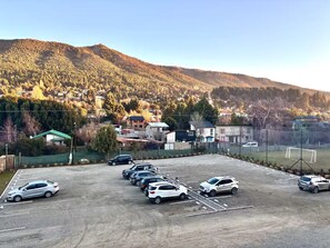 Un parking contenant plusieurs voitures, entouré d’une zone délimitée par une clôture, avec des maisons et une colline en arrière-plan.