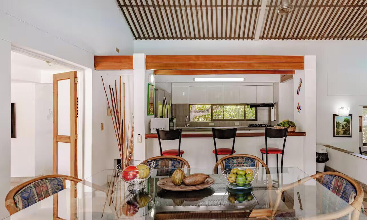 Un comedor con mesa de vidrio, sillas y cocina al fondo.