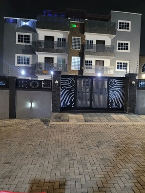 A multi-story residential building with a gated entrance, paved driveway, and a well-lit exterior. - your comfort is our priority (Accra)