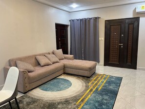 A living room with a beige sectional sofa, a white chair, a blue and gray rug, and a dark wooden door. - your comfort is our priority (Accra)