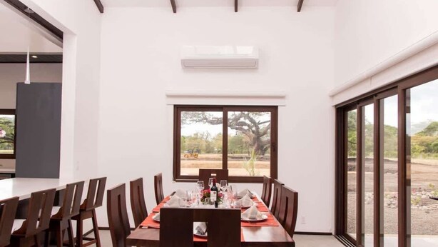 Comedor con una mesa larga dispuesta para seis, un ventanal con vistas al exterior y un equipo de aire acondicionado en la pared.