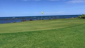 Campo de golf con un mástil de bandera en el centro, cielo despejado y el océano al fondo.