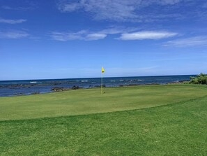 Ein Golfplatz mit Fahnenstange in der Mitte, blauem Himmel und dem Meer im Hintergrund.