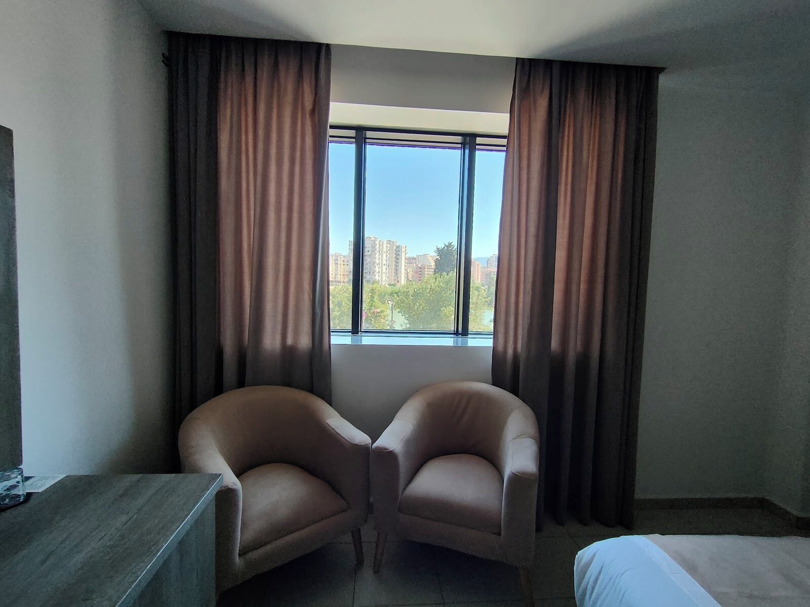 Une chambre d’hôtel avec une grande fenêtre, deux fauteuils, un lit et un bureau.