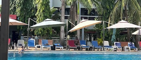 Un área de piscina con sillas de descanso y sombrillas, rodeada de palmeras y un edificio al fondo.