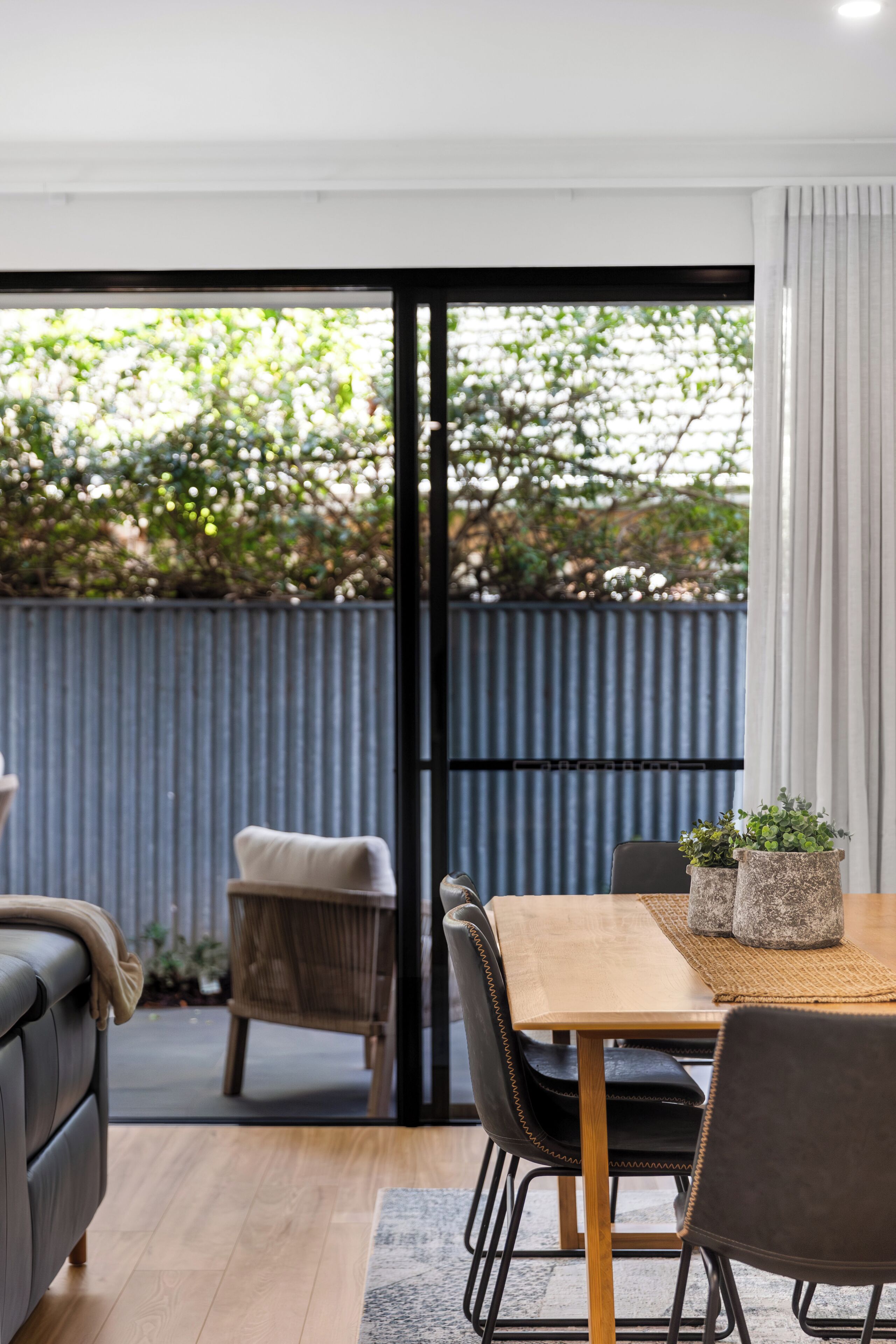 A modern dining area with a wooden table and chairs, a grey sofa, and a view of greenery through a glass door.