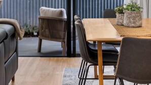 A modern dining area with a wooden table and chairs, a grey sofa, and a view of greenery through a glass door. - The Torrens A Contemporary Escape in the Foothills (Kingswood)