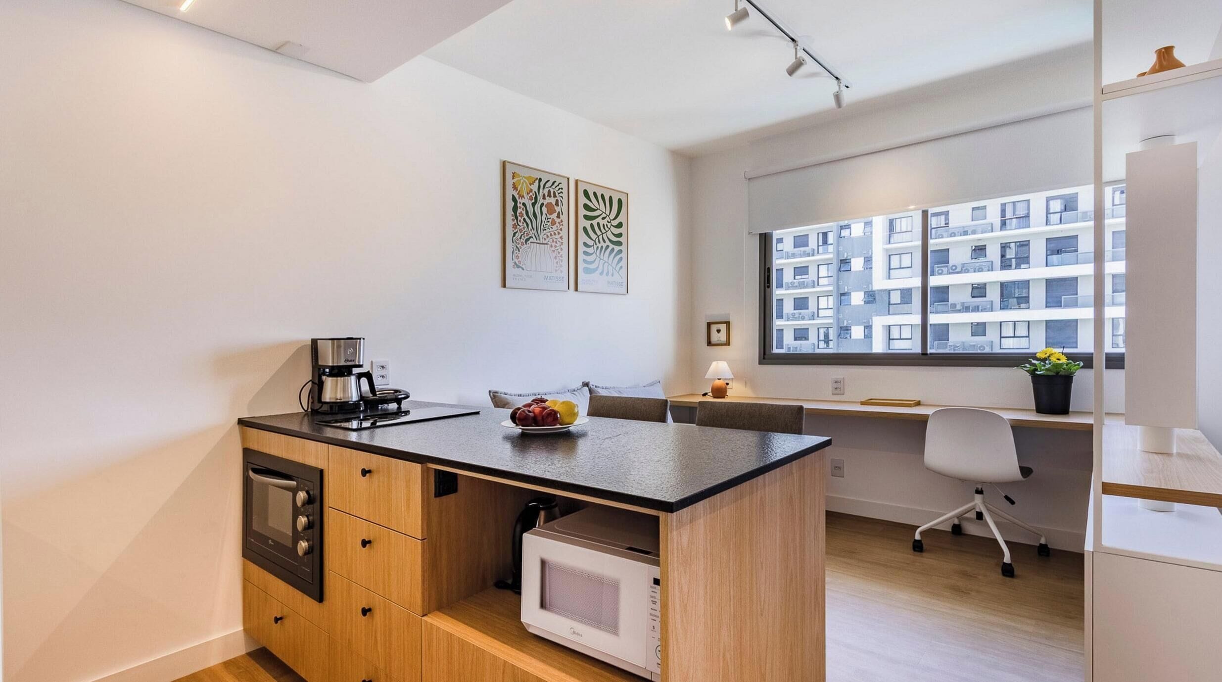 Una kitchenette compacta con cafetera, microondas y frutero. Se ve una ventana con vista a un edificio.