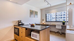 Una kitchenette compacta con cafetera, microondas y frutero. Se ve una ventana con vista a un edificio.