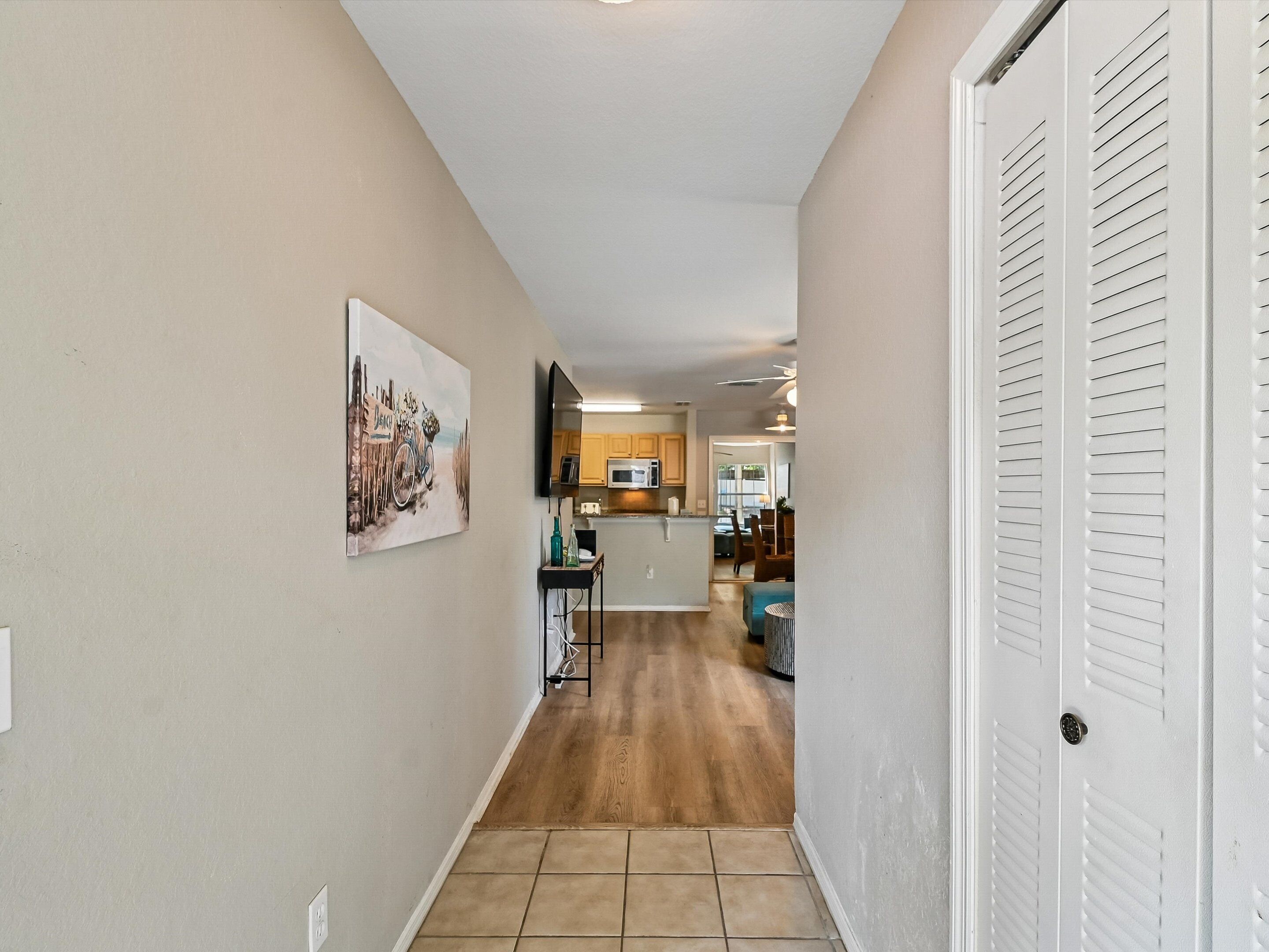 A hallway with a tiled floor, a painting on the wall, a kitchen area with wooden cabinets, and a white door with louvered panels.