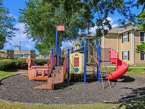 Ein Spielplatz mit verschiedenen Spielgeräten, umzäunt, mit Häusern im Hintergrund.