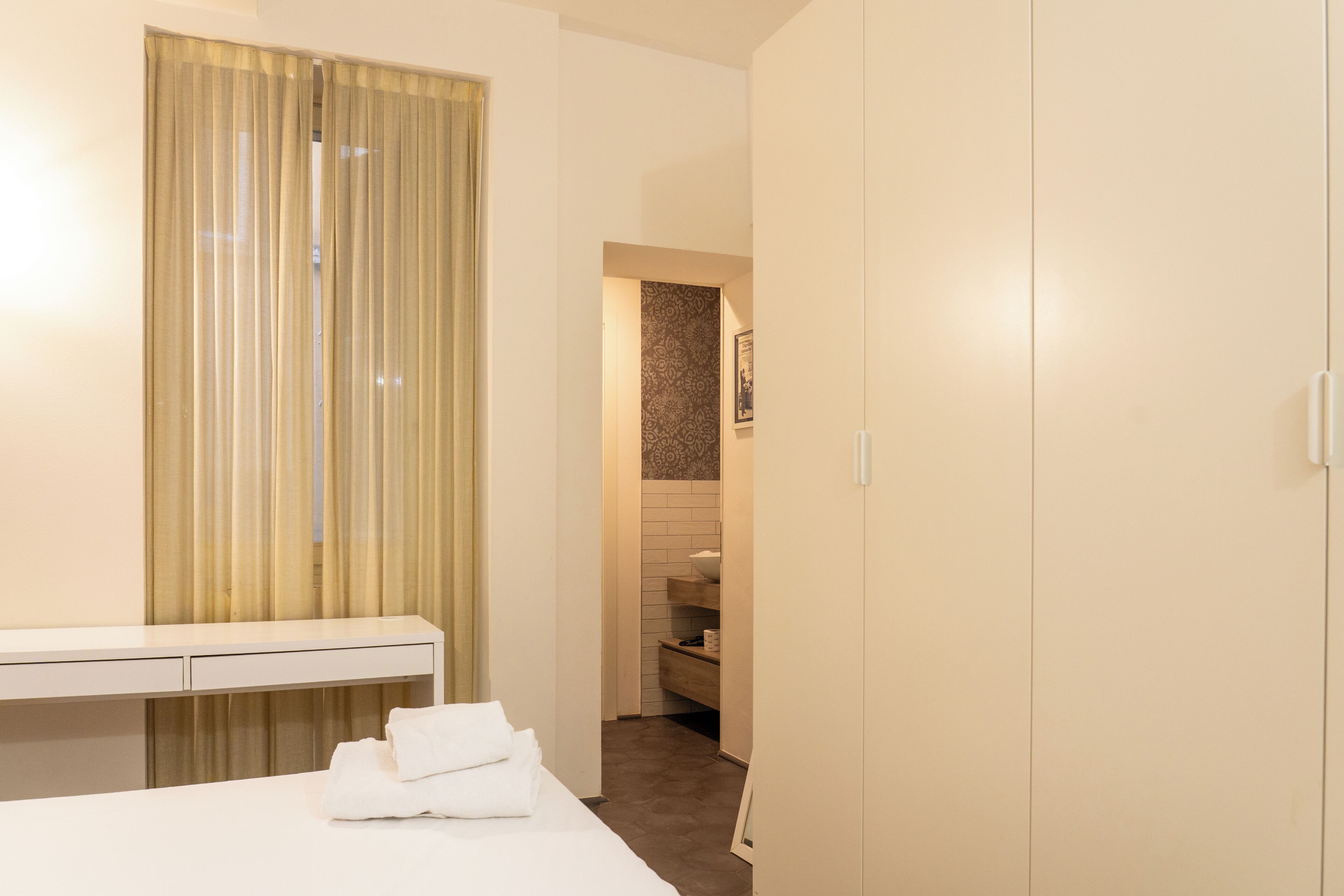 A hotel room with a bed, a desk, a wardrobe, and a bathroom visible through an open door.