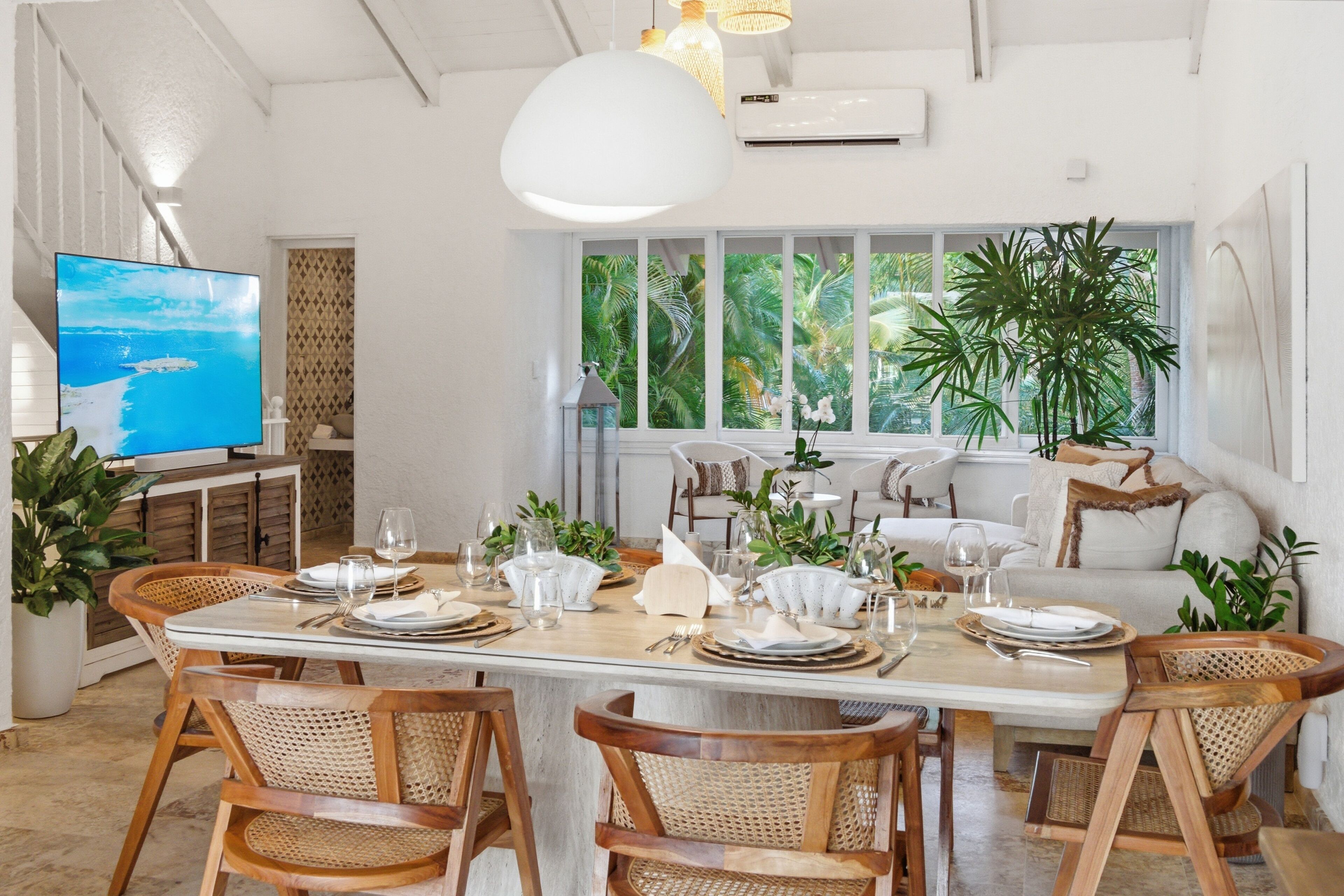 Un coin repas avec une longue table dressée pour six personnes, entourée de chaises en osier, et une grande fenêtre offrant une vue sur la végétation tropicale.