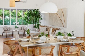 A dining area with a large table set for six, surrounded by wicker chairs, with a view of greenery through large windows.