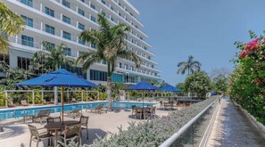 Área de piscina de hotel com várias espreguiçadeiras e guarda-sóis, céu claro e um edifício com varandas.